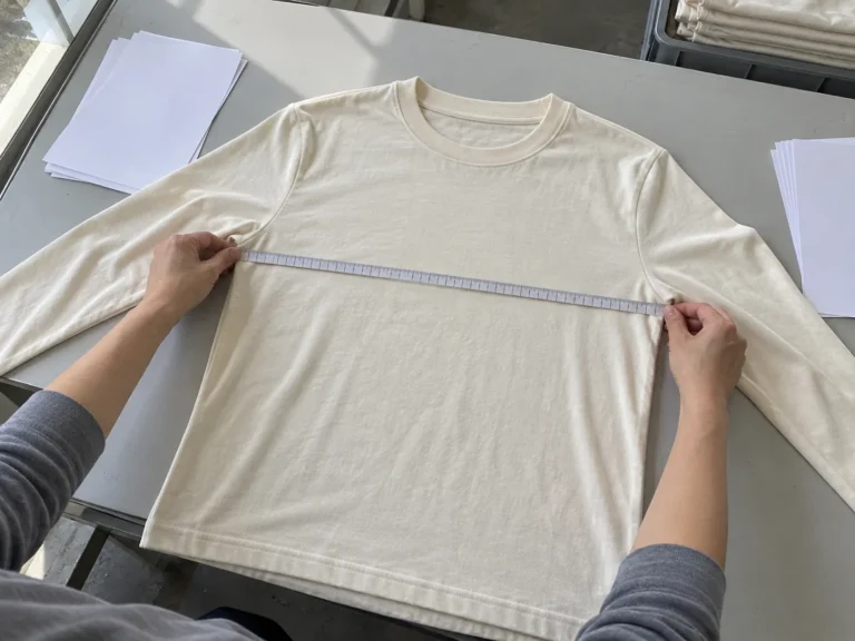 Photorealistic apparel QC documentary photograph, finished unbranded cream jersey long-sleeve laid flat on an inspection
